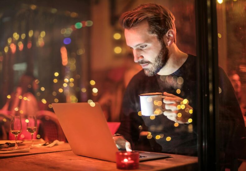 A young man with a beard is working on a laptop at a cozy cafe with warm lighting, holding a coffee cup.